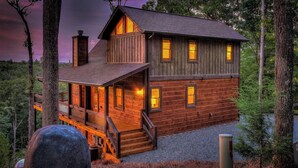 A two-story wooden cabin with a porch and multiple windows, surrounded by trees. - Songbird Ridge - Aska Adventure Area | Panoramic Views | Hot Tub | Outdoor Fireplace (Blue Ridge)