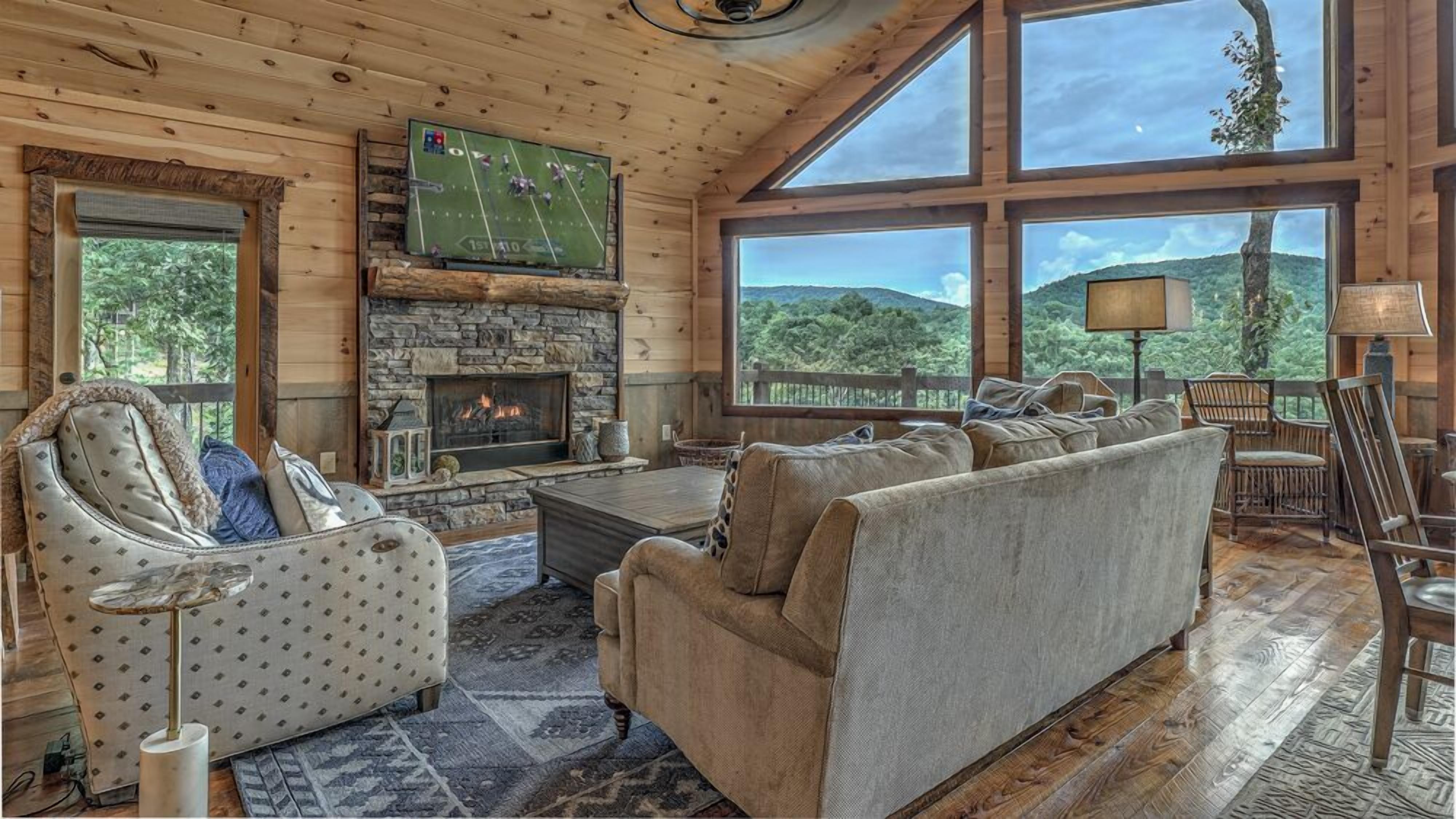 A cozy cabin interior with a stone fireplace, a large window offering a scenic view, and a comfortable seating area.