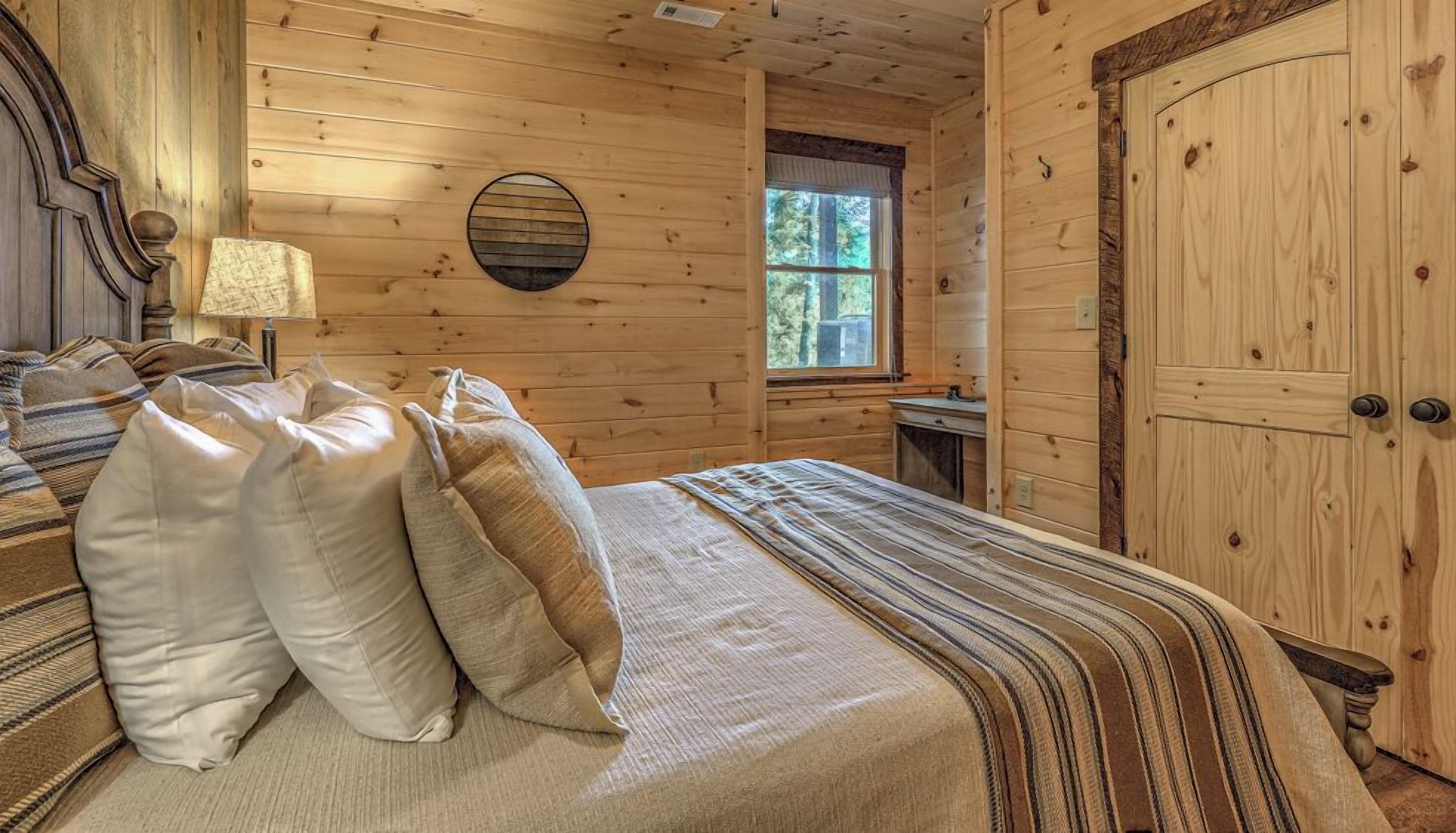 Un dormitorio de cabaña de madera con una cama, almohadas, un espejo redondo y una ventana.