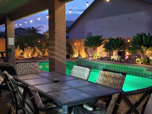 Un patio al aire libre con piscina, rodeado de palmeras y luces colgantes.