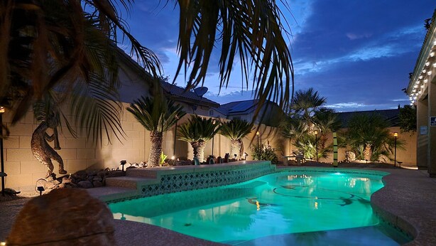 A swimming pool with a tiled deck, surrounded by palm trees and a building with a tiled roof.