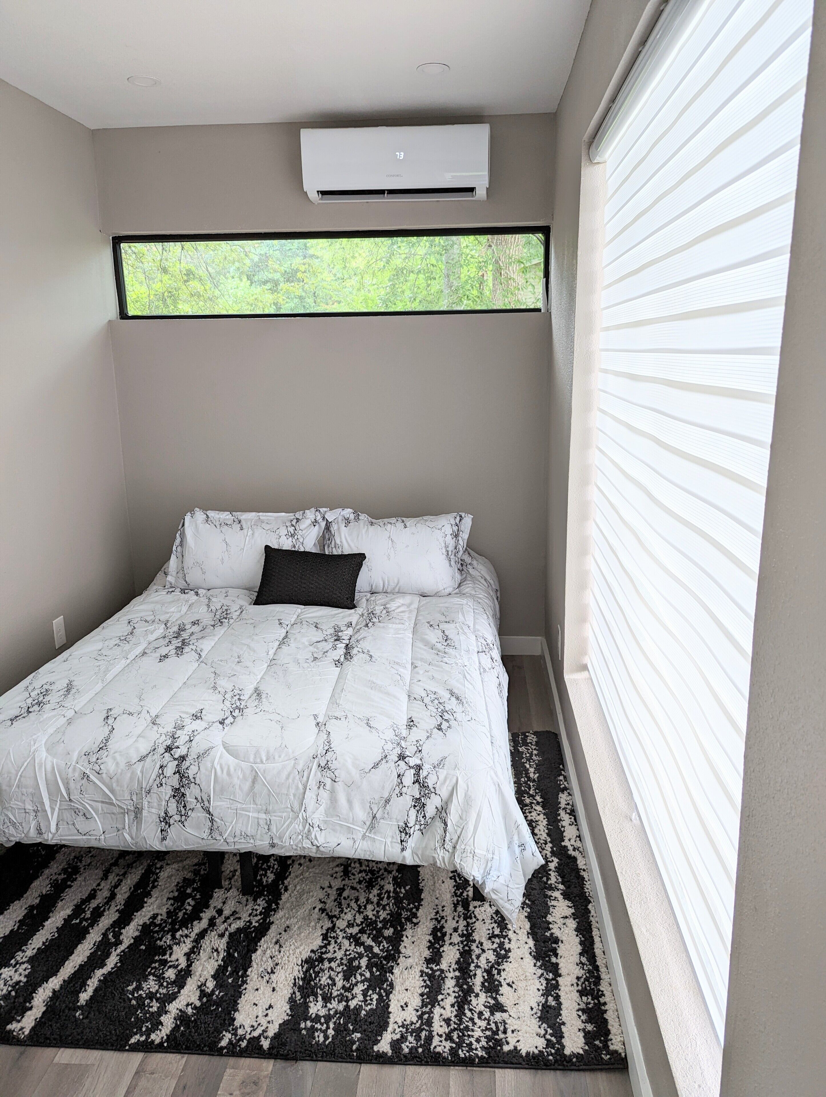 A bedroom with a bed, a window with blinds, an air conditioning unit, and a patterned rug.