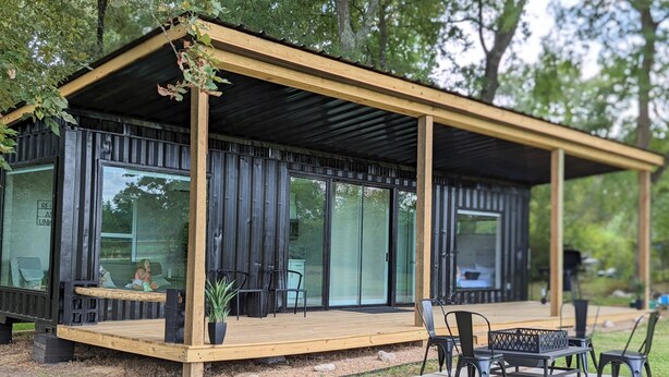 Una cabaña moderna y compacta con terraza de madera, ventanas amplias y mobiliario exterior.