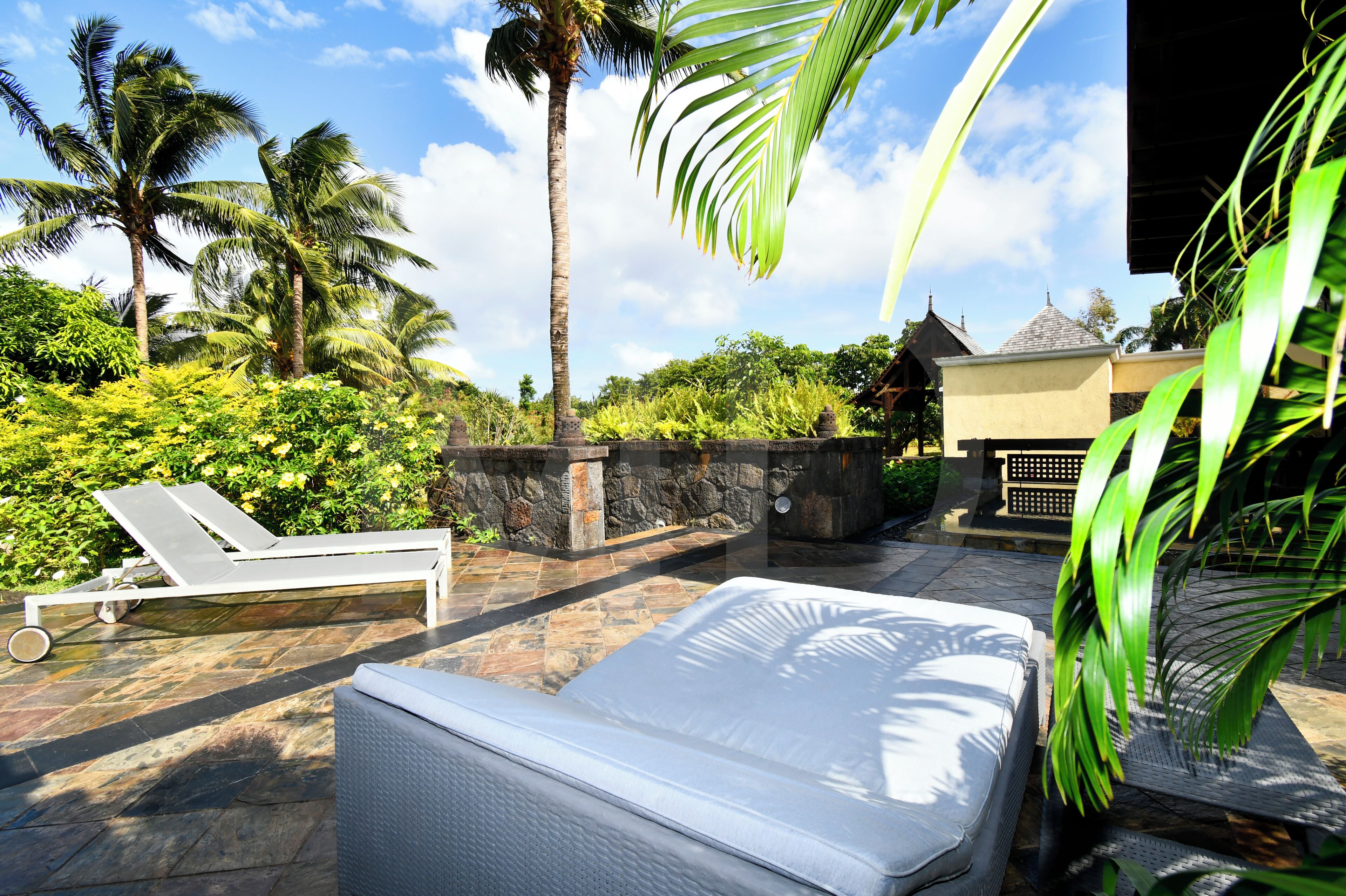 A sunlit outdoor area with a lounge chair, a mattress, and a stone wall.