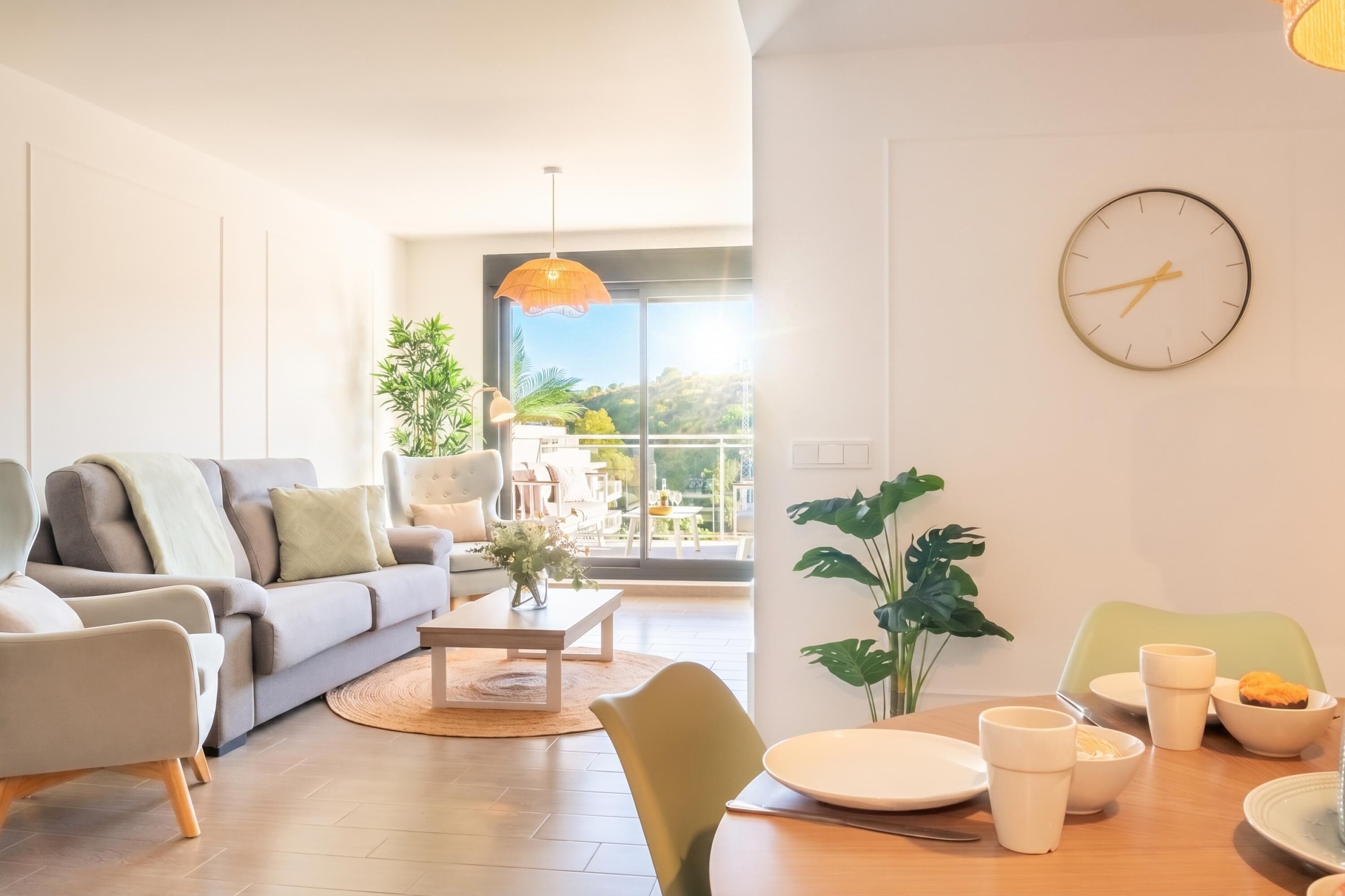 Ein modernes Wohnzimmer mit einer Couch, Sesseln, einem Esstisch mit Tassen und Tellern, einer Uhr und einem großen Fenster mit Aussicht.