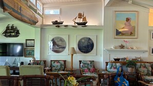 Une pièce spacieuse avec un plafond haut, décorée sur le thème de la marine, avec des maquettes de navires, des tableaux représentant des paysages marins et une cheminée.