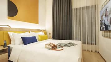 Habitación de hotel con una cama grande, una mesita de noche con lámpara, un televisor montado en la pared y una ventana con cortinas.
