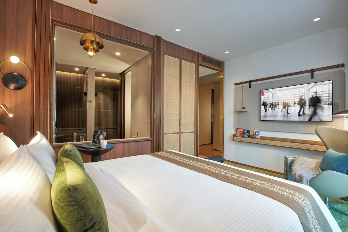 Ein Hotelzimmer mit einem großen Bett, einer Sitzecke mit Sessel, einem kleinen Tisch, einem Spiegel und einem an der Wand montierten Fernseher.