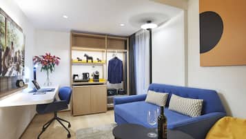 A modern hotel room with a desk, laptop, chair, sofa, and a small table with a wine glass.