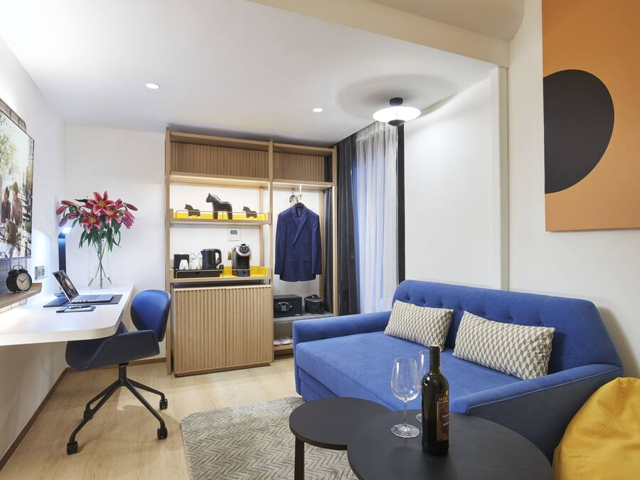 A modern hotel room with a desk, laptop, chair, sofa, and a small table with a wine glass.