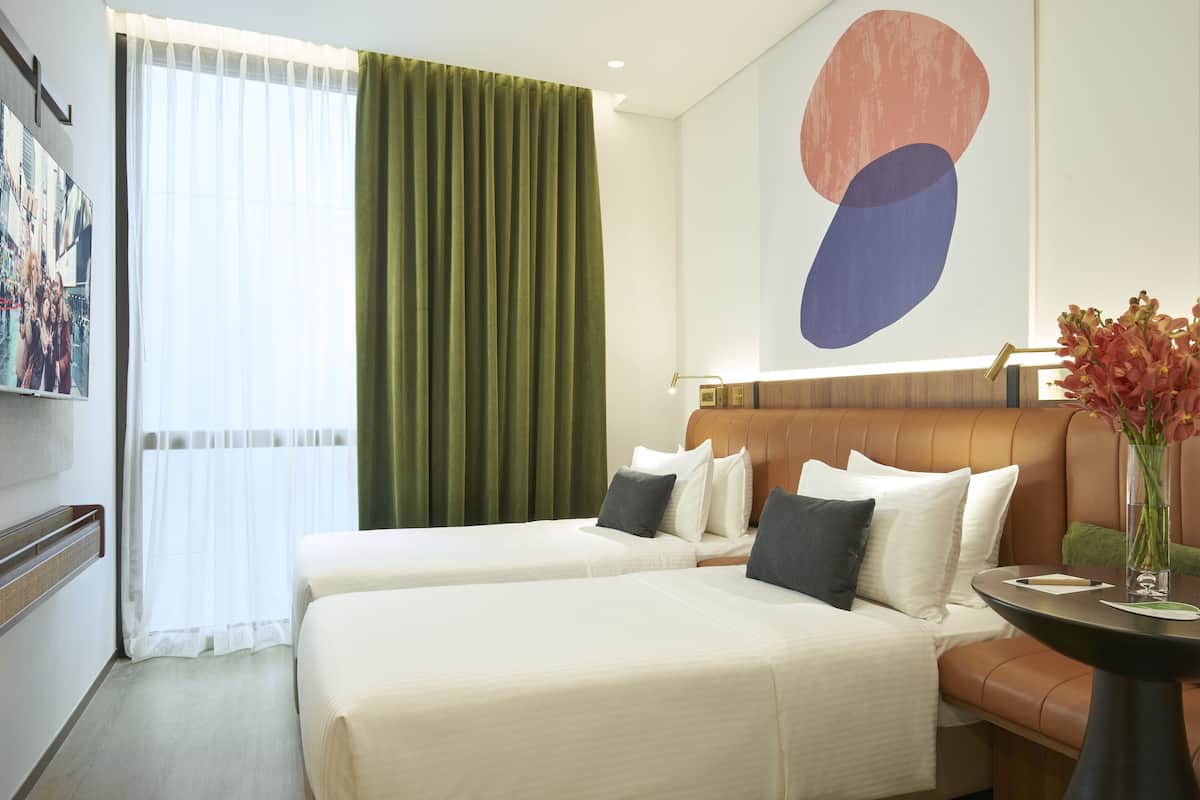 Ein Hotelzimmer mit zwei Betten, einem grünen Vorhang, einem modernen abstrakten Gemälde und einer Vase mit Blumen auf einem Tisch.