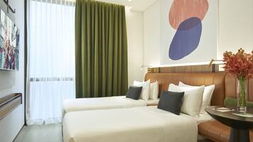 A hotel room with two beds, a green curtain, a modern abstract painting, and a vase with flowers on a table.