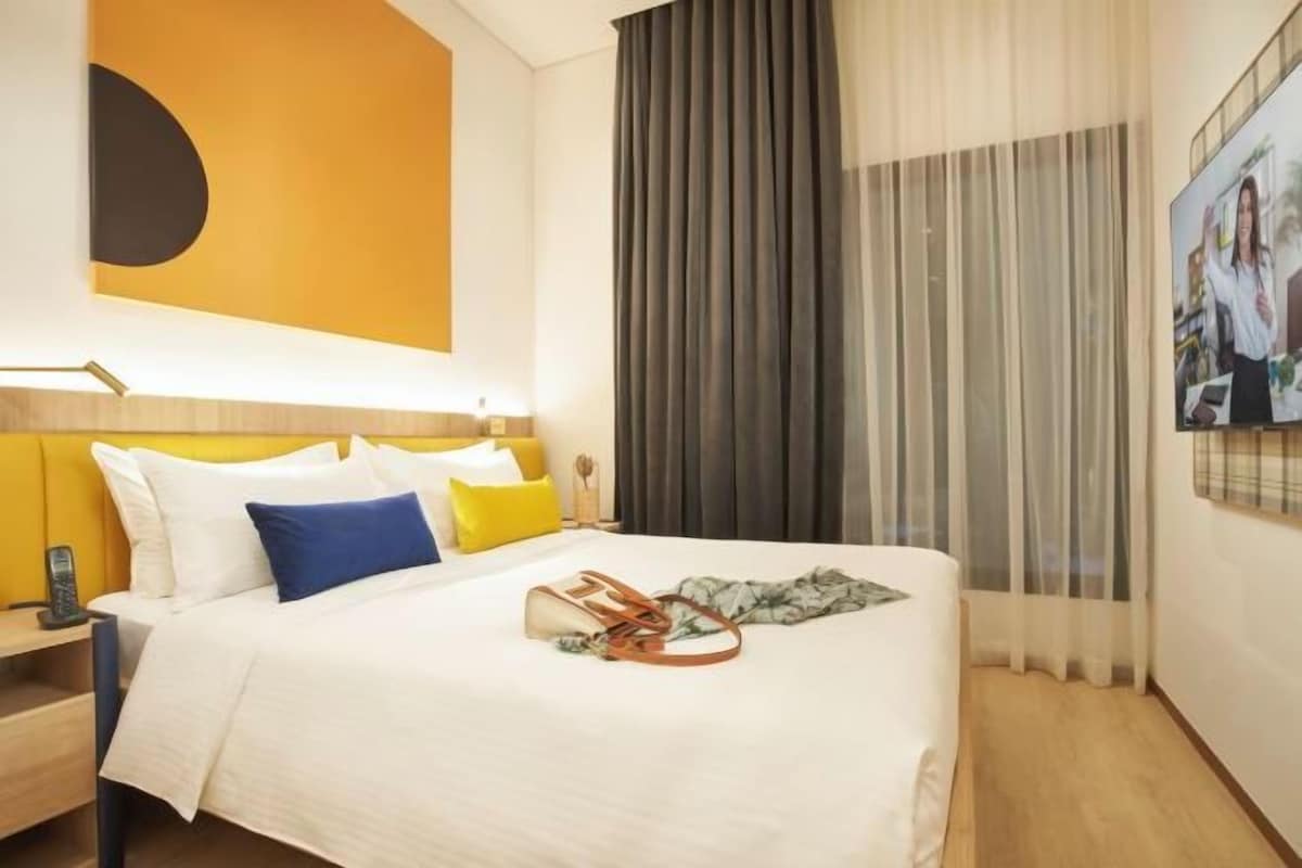 Habitación de hotel con cama, mesita de noche, televisor montado en la pared y una ventana grande con cortinas.