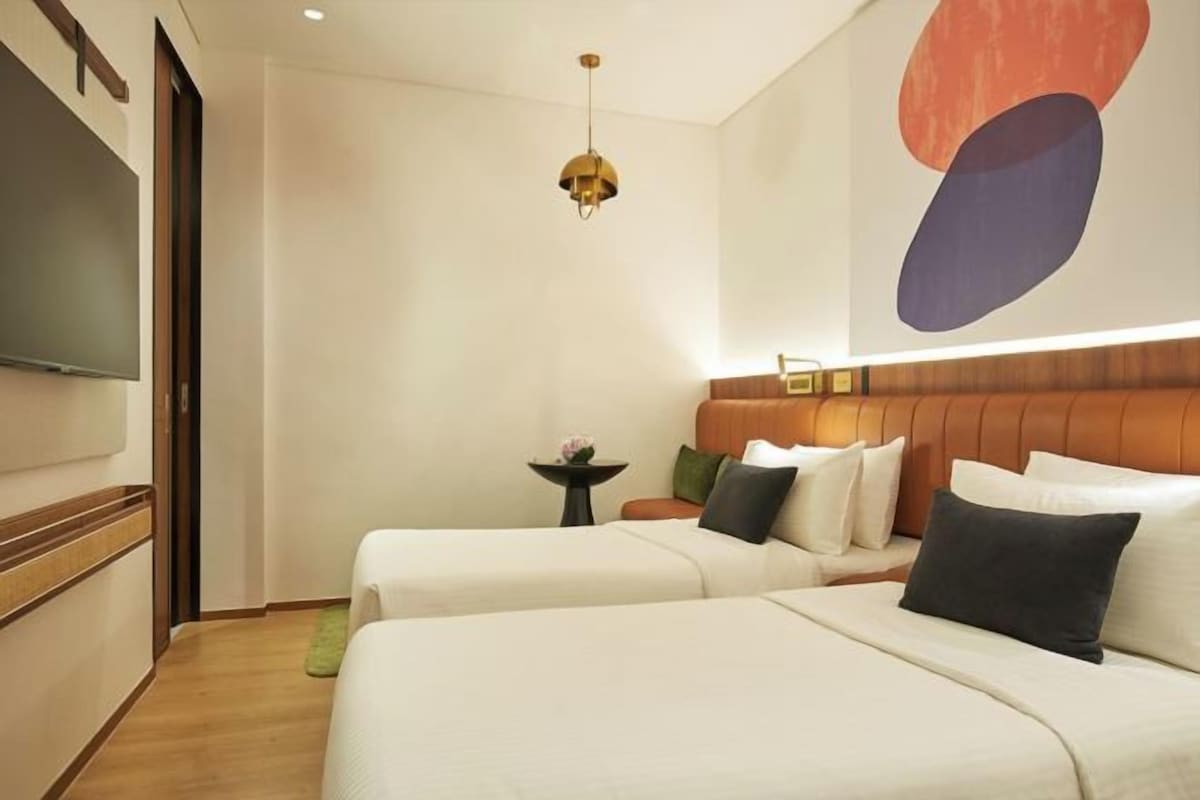 Habitación de hotel con dos camas, un televisor, una mesita con una flor y una obra de arte en la pared.