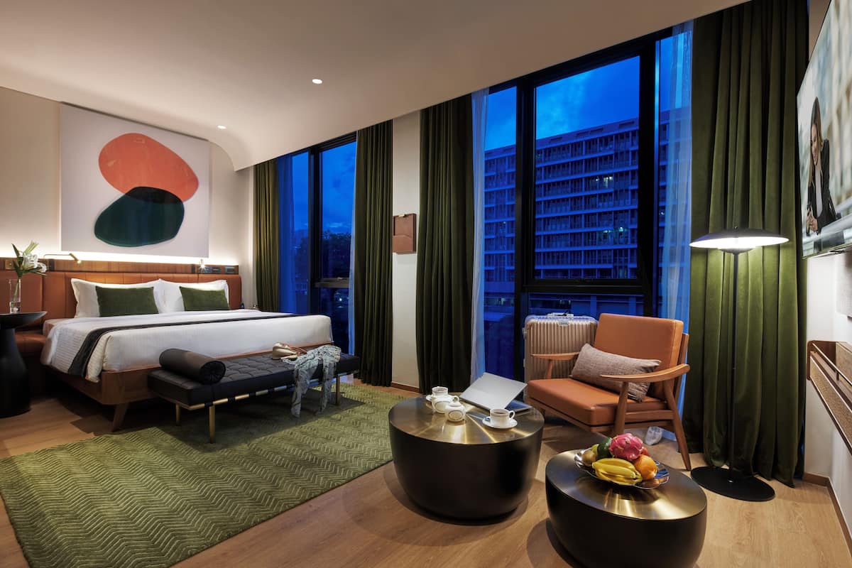 Ein modernes Hotelzimmer mit einem großen Bett, einer Sitzecke mit Couchtisch und Blick auf eine Stadtkulisse durch große Fenster.