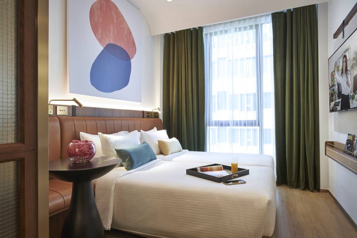 Ein Hotelzimmer mit einem großen Bett, einem Nachttisch mit einer Vase und Blick auf die Stadt durch das Fenster.