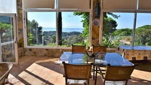 A dining area with a glass table and wicker chairs, large windows with a view of the sea and greenery, and stone walls.