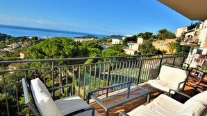 A balcony with two white cushioned chairs and a small table, overlooking a coastal town with houses and greenery.