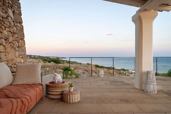 Relax in a serene outdoor seating area with a sofa, wicker tables, and a potted plant, overlooking the sea and coastline.