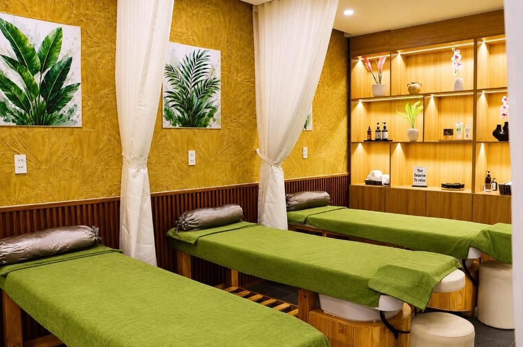 Una habitación con tres camillas de masaje, cada una con una cubierta verde y un cojín. Hay dos cuadros grandes de plantas en la pared, y una estantería con varios objetos, incluyendo botellas y un cartel que dice 'Te mereces relajarte'.