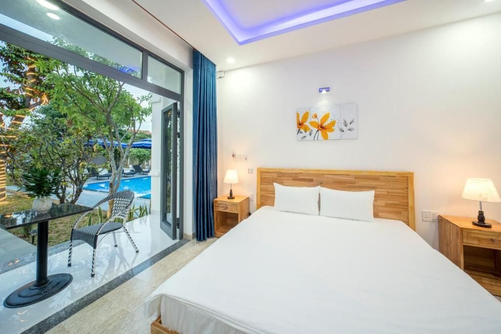 A modern hotel room with a large bed, two bedside tables, a view of a swimming pool, and a balcony with outdoor furniture.