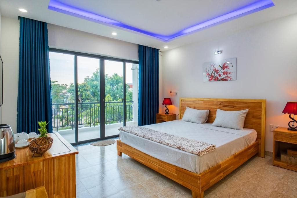 A hotel room with a large bed, a wooden nightstand, a desk with a kettle, and a view of greenery through the balcony doors.