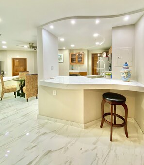 Una cocina moderna con una isla curva, una zona de comedor con sillas de mimbre y un suelo de mármol.