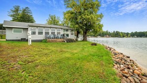 Uma cabana à beira do lago com varanda e mobiliário para áreas externas, rodeada por um gramado e um muro de pedra.