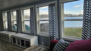 Quarto com vista para um lago, amplas janelas com cortinas em padrão geométrico, um banco e um sofá com almofadas estampadas.