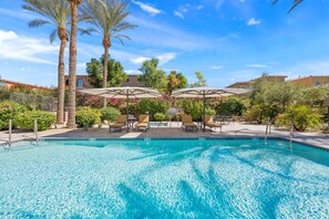 A clear swimming pool with lounge chairs and umbrellas, surrounded by palm trees and a clear sky.