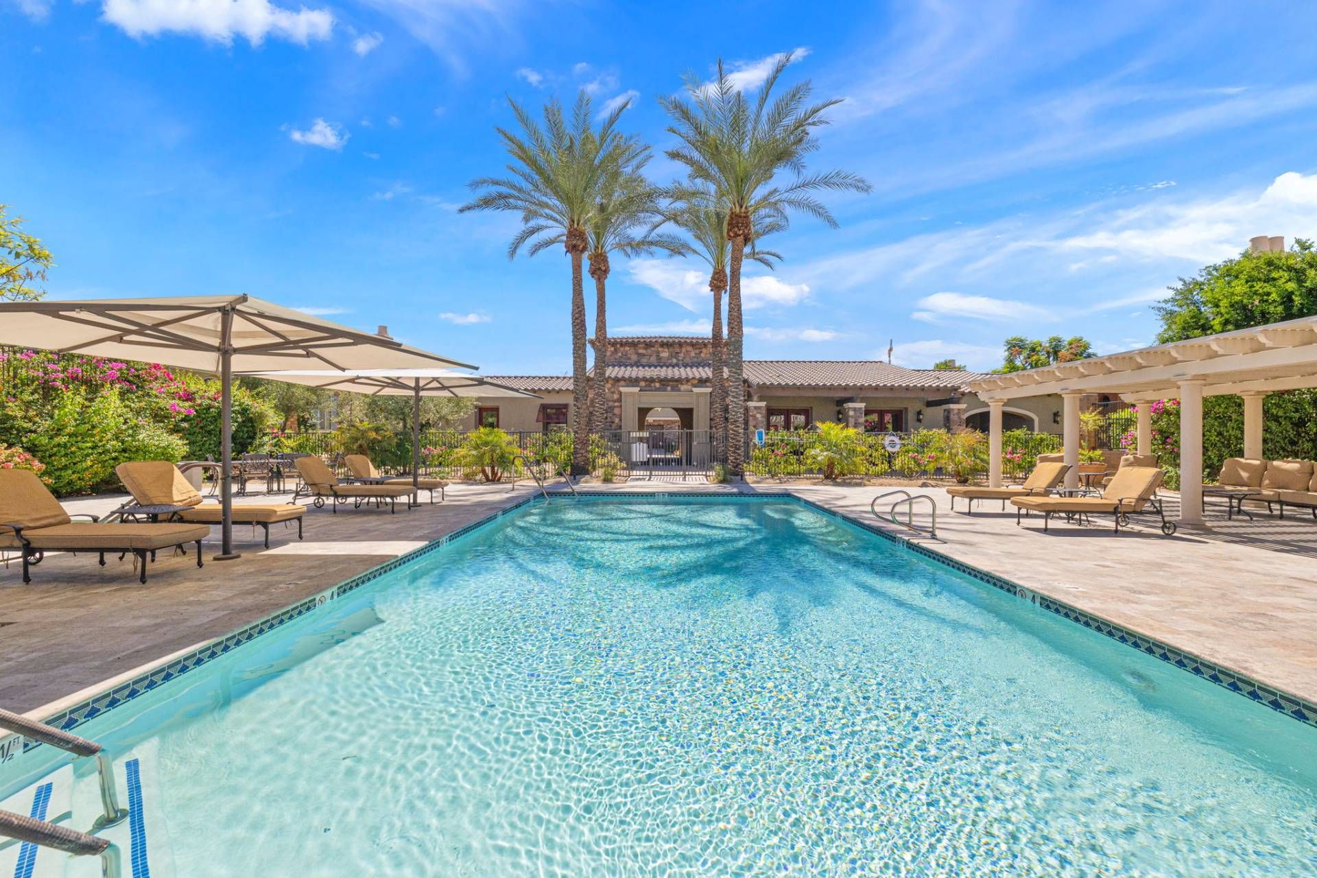 A swimming pool with lounge chairs and umbrellas, surrounded by palm trees and a building with a tiled roof.