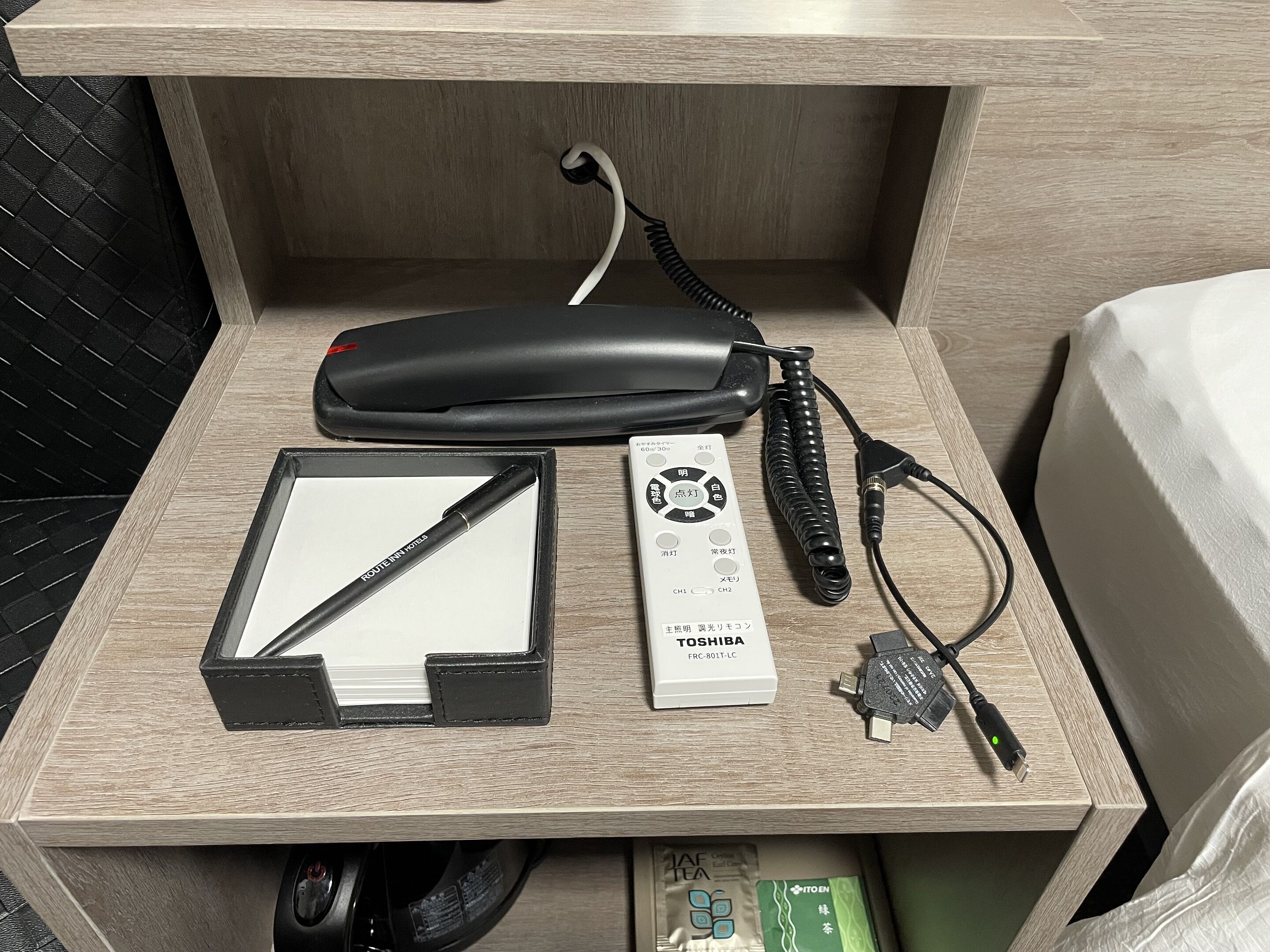 A hotel room with a wooden desk, a black telephone, a white remote control, a black pen, a power strip, and a clock.