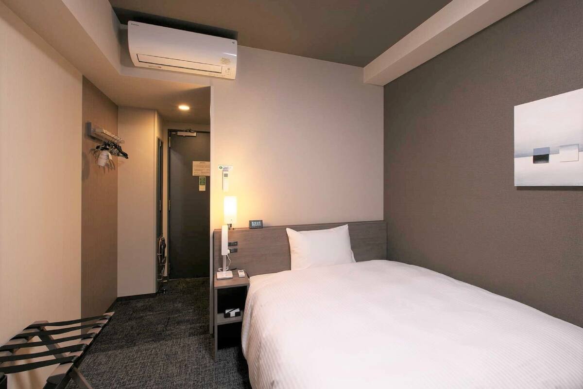 Una habitación de hotel con cama, mesita de noche, lámpara y aire acondicionado.