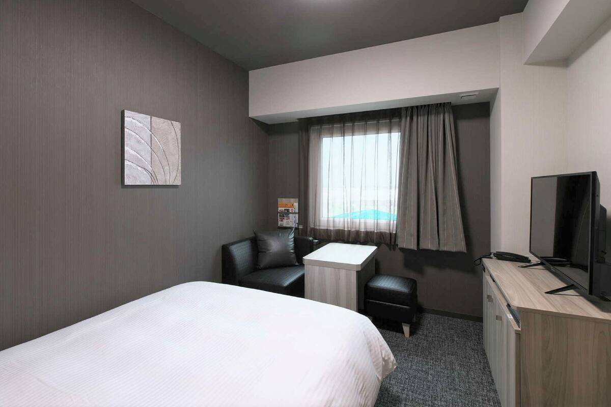 Habitación de hotel con cama, escritorio, silla, televisor y ventana con cortinas.