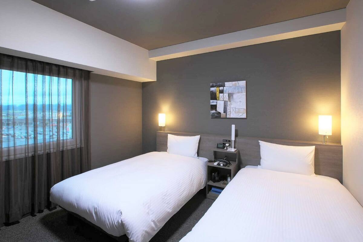 Habitación de hotel con dos camas, una mesita de noche, una lámpara, un cuadro en la pared y una ventana con cortinas.
