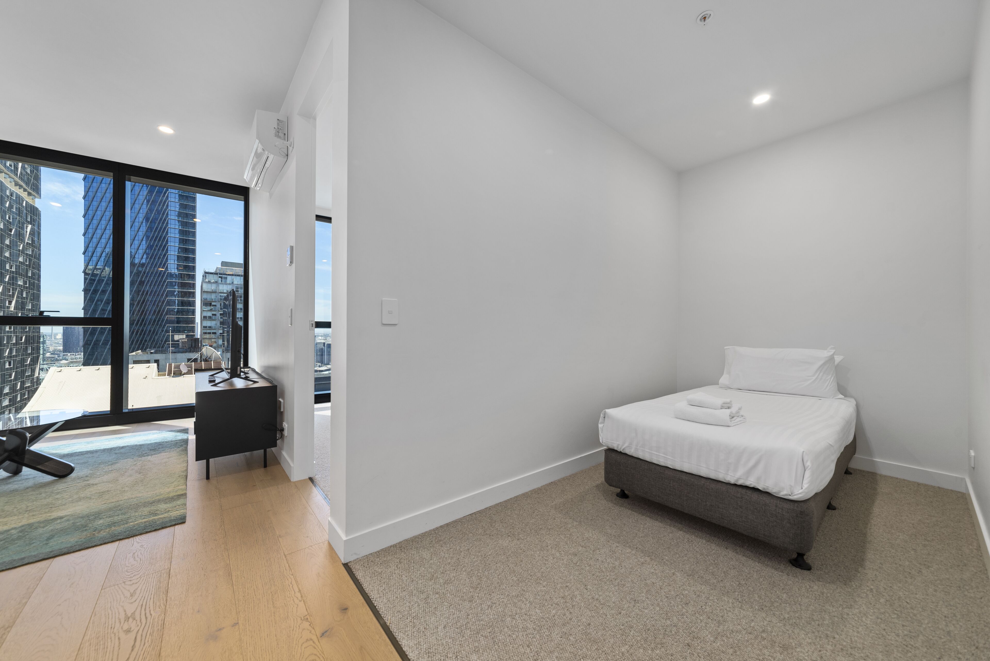A modern bedroom with a large window, a bed, a desk, and a view of the cityscape.