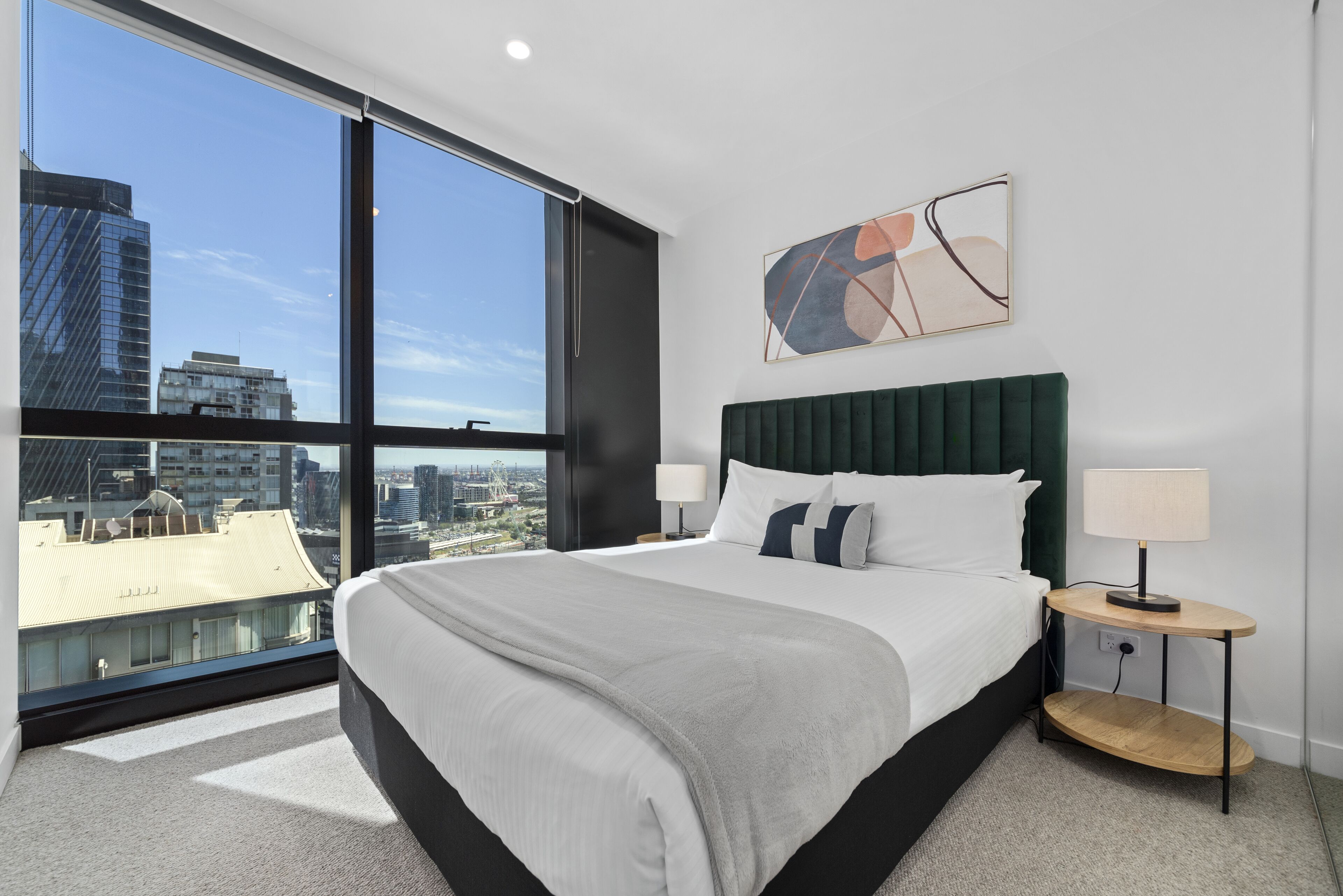 Una camera d'albergo moderna con un letto grande, un comodino, una lampada e vista sulla città attraverso ampie finestre.