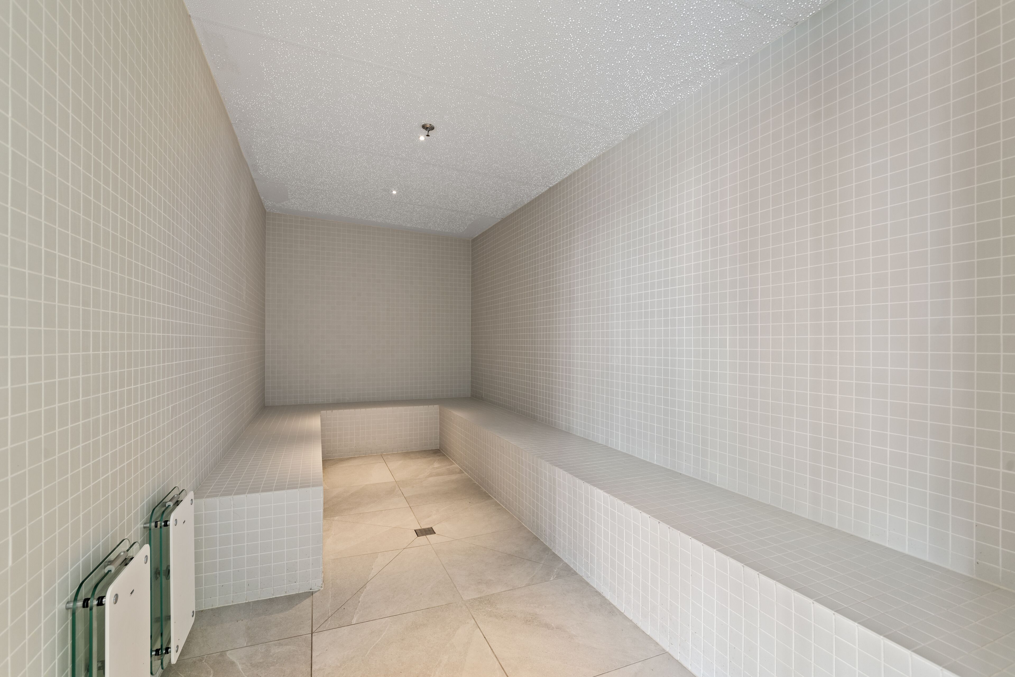 A bathroom with white tiled walls and floor, a bench, and a glass partition.
