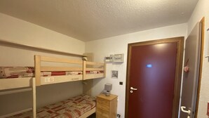 A small, compact room with bunk beds, a wooden dresser, a door, and a small table with a lamp. - Studio for 4 near shops, 450 m to slopes (Les Carroz d'Arâches)