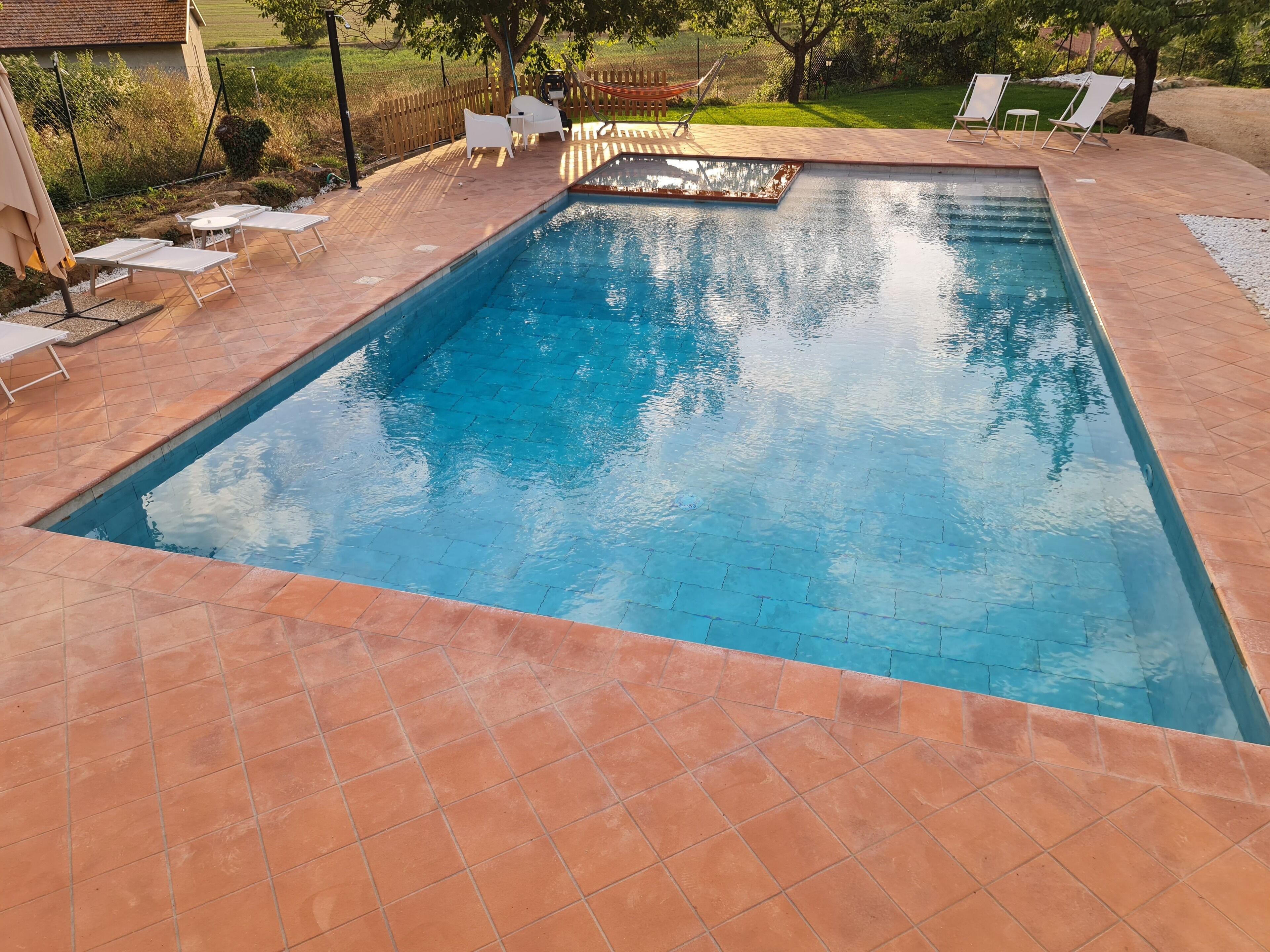 A rectangular swimming pool with a tiled deck, surrounded by white lounge chairs and a hammock.