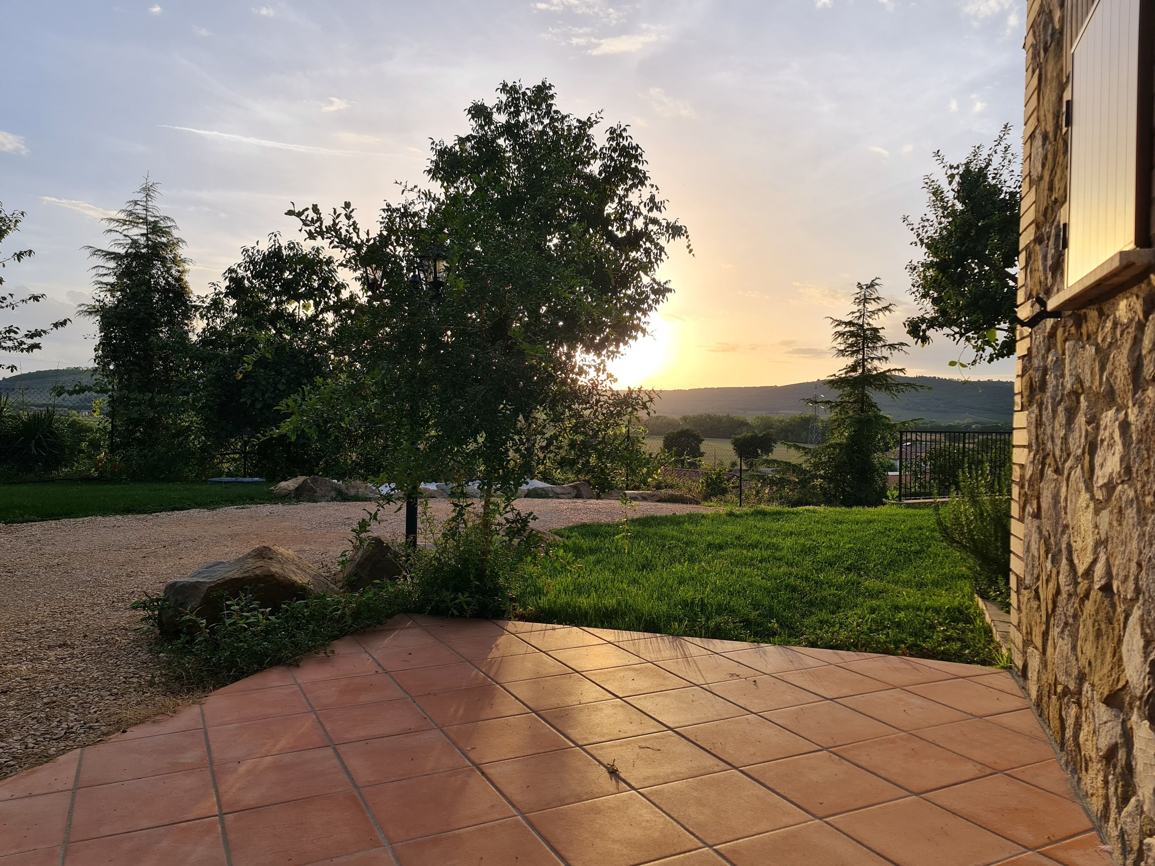Une terrasse avec un mur en pierre, donnant sur un champ verdoyant et des arbres, au coucher du soleil.