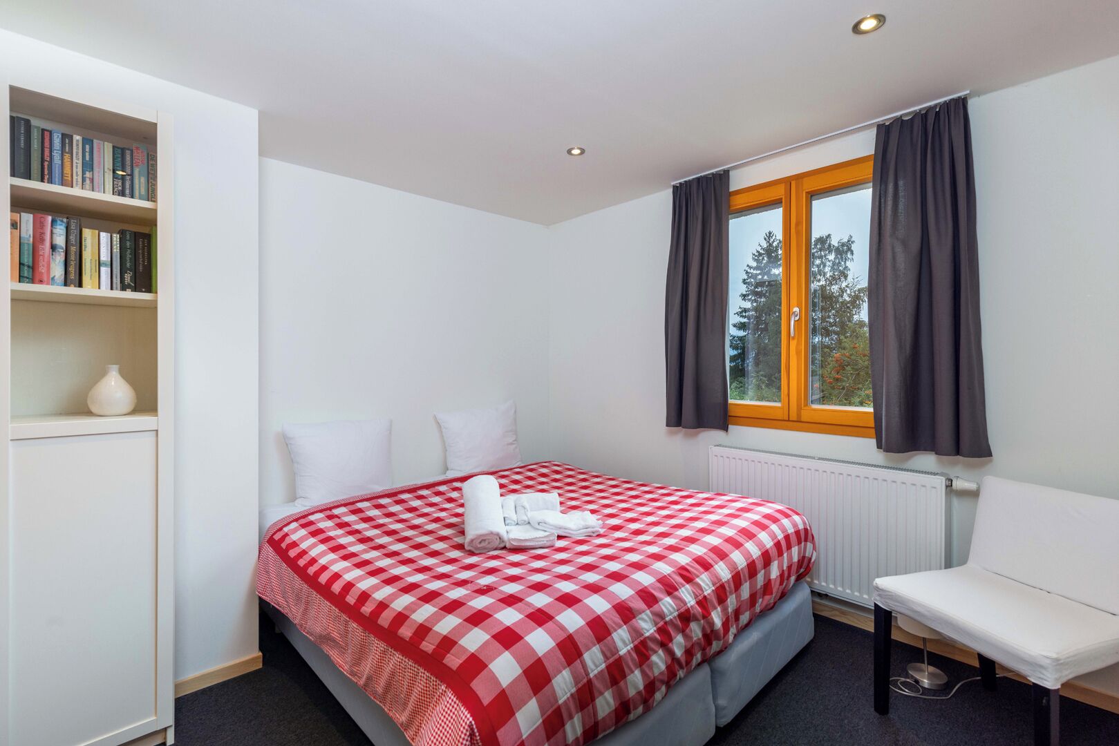 A bedroom with a checkered bedspread, a white chair, a bookshelf, and a window with curtains.