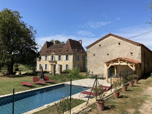 Una casa de piedra con piscina, rodeada por un jardín vallado con plantas en macetas y sillas reclinables rojas.