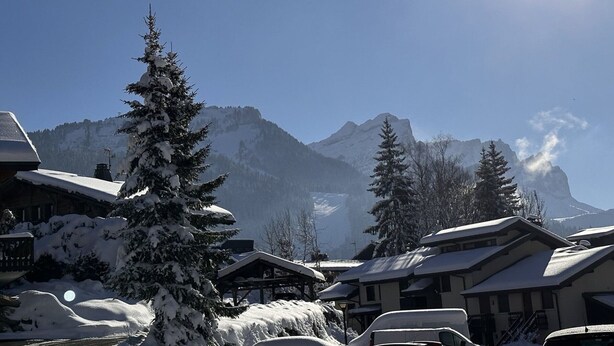 A snowy mountain village with chalet-style buildings, snow-covered trees, and a clear view of the mountains in the background.