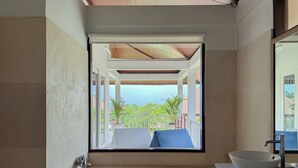 Ein Badezimmer mit einer Badewanne, einem Waschbecken mit weißer Schüssel und Blick auf einen Balkon mit Geländer und Grünpflanzen.