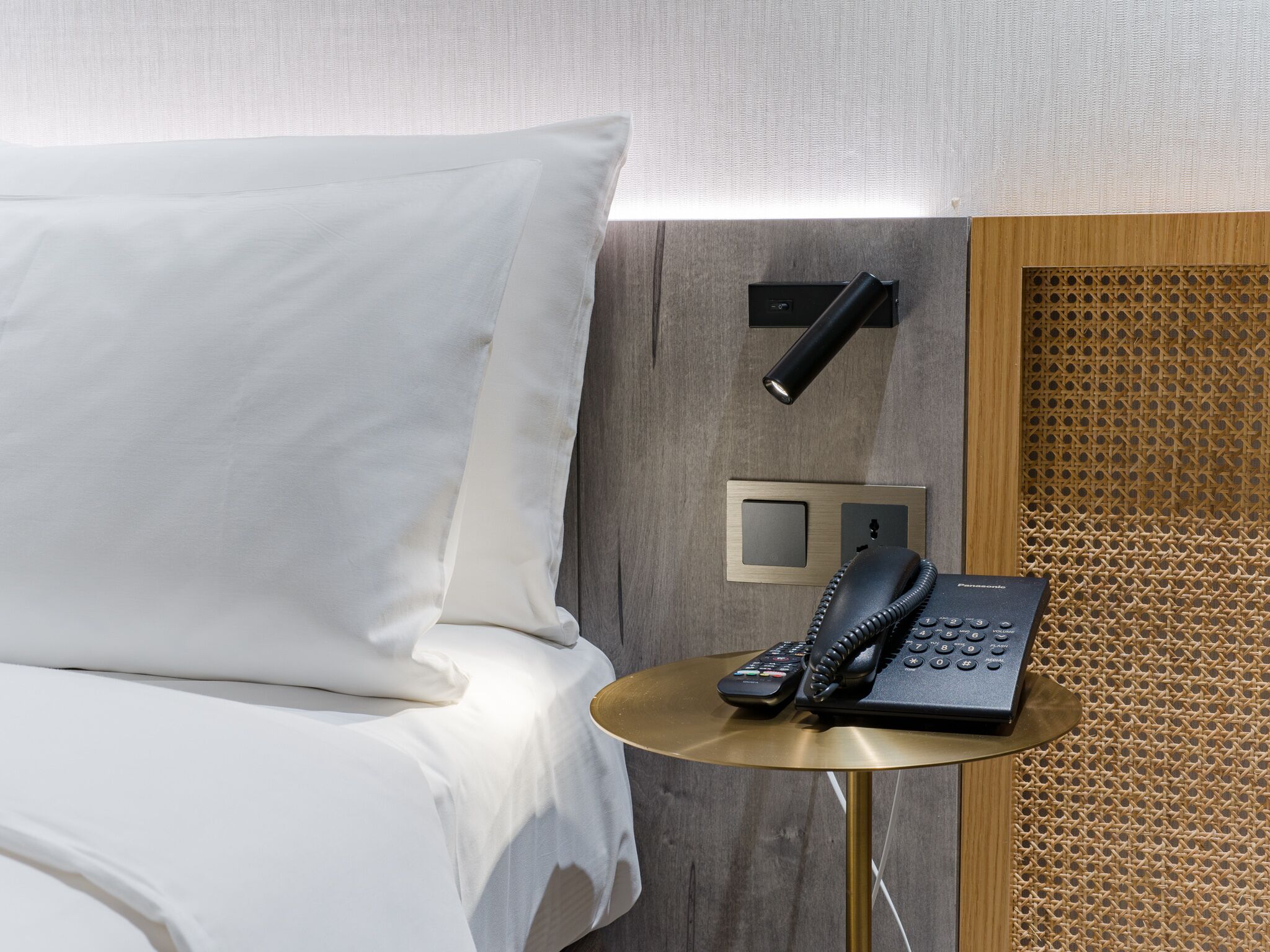 A hotel room with a bed, a bedside table with a telephone, and a wall-mounted light.