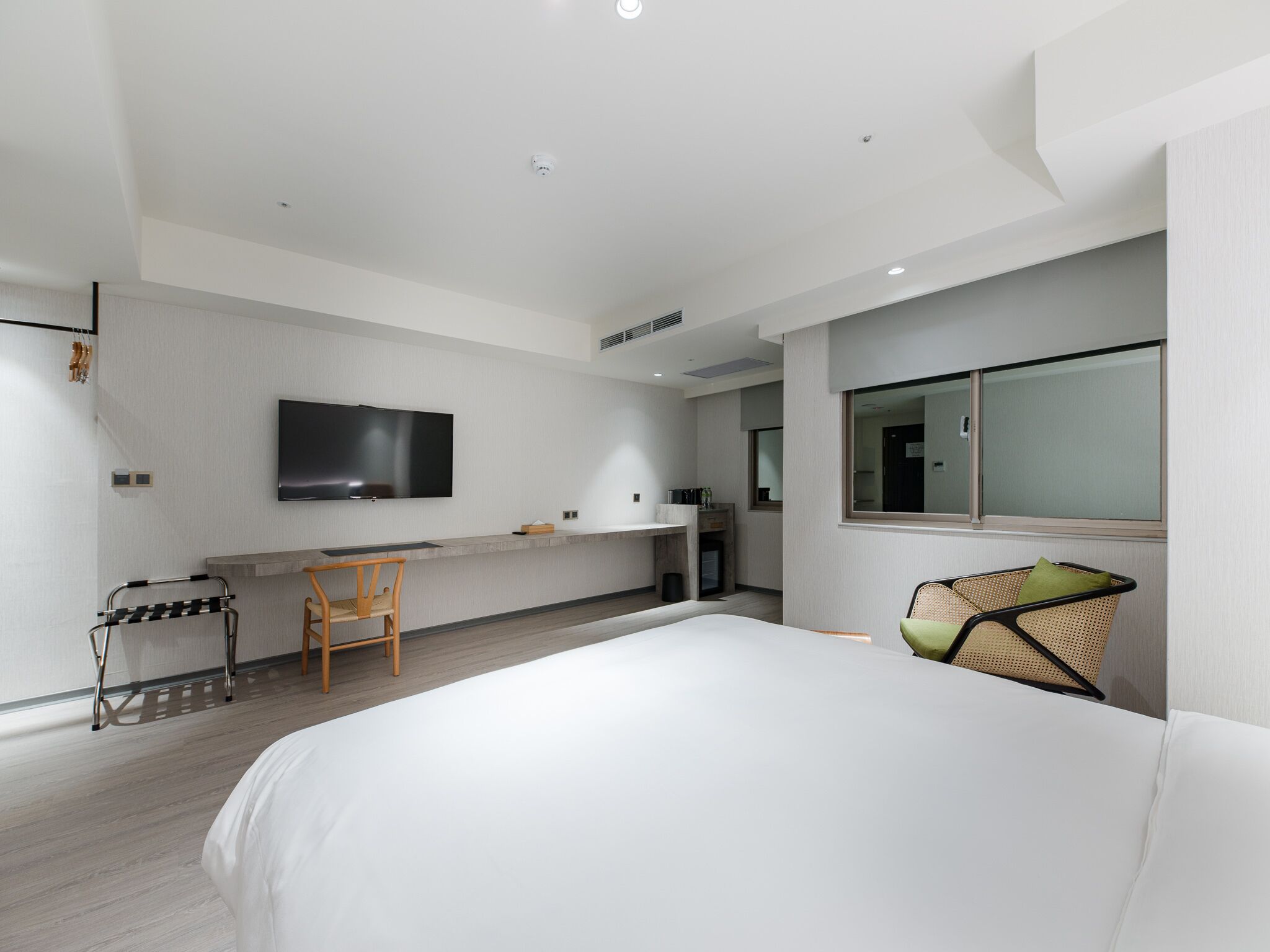 A modern hotel room with a large bed, a desk with a chair, a flat-screen TV mounted on the wall, and a window with a view.