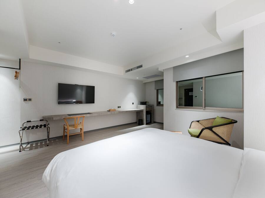 A modern hotel room with a large bed, a desk with a chair, a flat-screen TV mounted on the wall, and a window with a view.