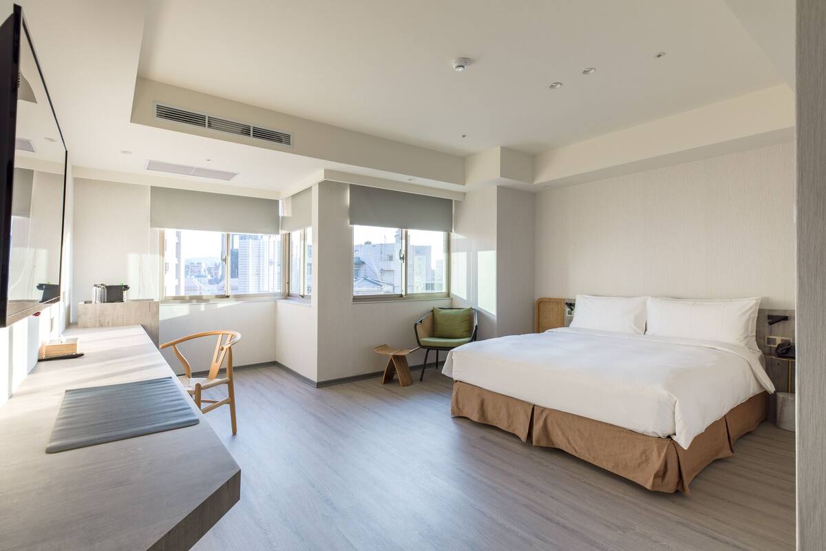 A modern hotel room with a large bed, a desk, a chair, and a view of the city.