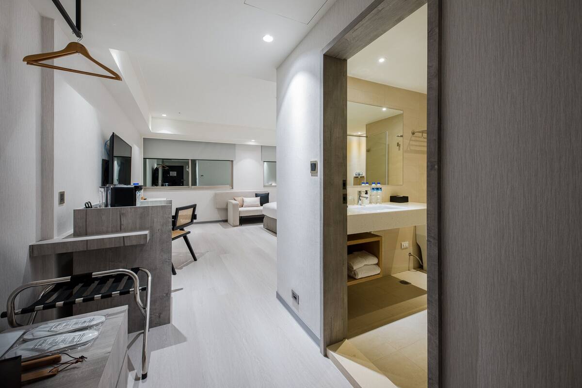A modern hotel room with a large bed, a desk, a chair, a TV, and a bathroom with a sink and mirror.